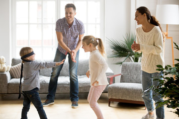 Wall Mural - Blindfolded cute little boy playing hide and seek at home, parents and kids laughing spending time together enjoying game on weekend, happy family of four having fun leisure activity in living room