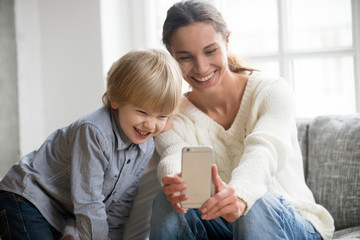 Wall Mural - Happy mother laughing taking selfie with little son on smartphone at home, smiling single mom and cute adopted boy playing making photo posing for self portrait, mommy and kid watching video on cell