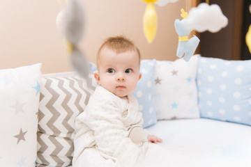 Cute baby sitting in a white round bed. Light nursery for young children.  Toys for infant cot. Smiling child playing with mobile of felt in sunny bedroom.