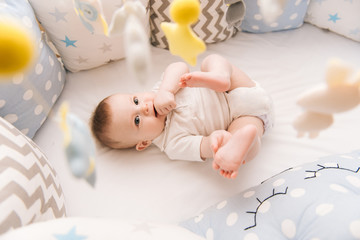 Wall Mural - Cute baby lies in a white round bed. Light nursery for young children.  Toys for infant cot. Smiling child playing with mobile of felt and chewing his feet in sunny bedroom.