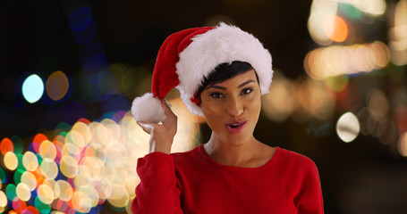 Wall Mural - Cheerful young woman modeling Santa hat in outdoors setting with bokeh lights