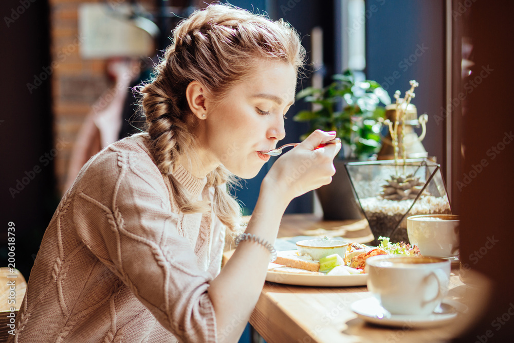 Charming woman with cute hairstyle having lunch during the rest in coffee shop, happy Caucasian female eating breakfast while relaxing in cafe during free time - obrazy, fototapety, plakaty 