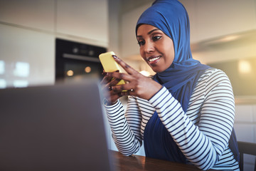 Wall Mural - Smiling Arabic entrepreneur sitting in her kitchen sending text