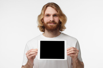 Positive bearded man isolated on grey background standing in casual clothes holding tablet and showing it blank screen with happy smile as if advising product or service