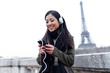 © nenetus - Smiling asian young woman listening to music with mobile phone in front of Eiffel tower in Paris.