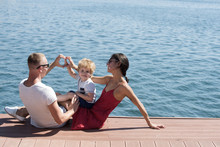 Mother And Father Making Heart Or Love Gesture With Hands Near Their Child. Happy Family Spend Time Together, Sea Background. Parents With Son Sit On Seafront, Rear View. Family Vacation Concept.