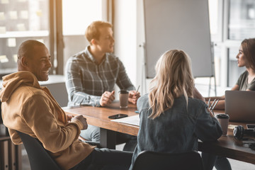 Poster - Four young people sitting at the office desk together. They conversing and laughing. Gadget and coffee cups are on desk