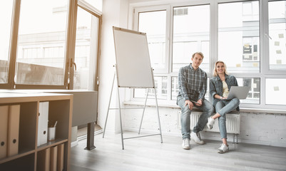 Aufkleber - Portrait of man and woman sitting near office window and smiling at camera. They keeping gadgets in hands. Copy space in left side
