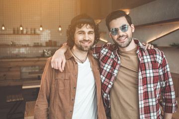 Best friends. Waist up portrait of happy two young men standing and embracing in cafeteria. They are looking at camera and smiling