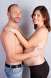 Handsome bald man and his beautiful pregnant wife are hugging and smiling while standing near wall at home
