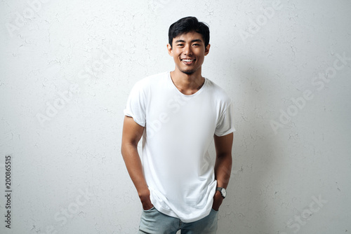 Foto Smiling asian guy in white blank t-shirt, grunge wall, studio portrait