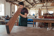 © mavoimages - Young woodworker examining a plank of wood in his workshop
