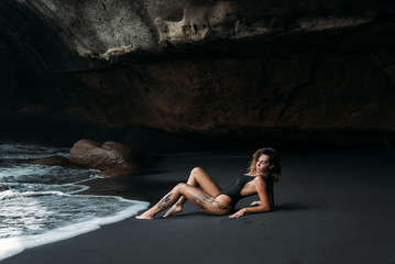 Wall Mural - Beautiful girl with sexy body in black swimsuit lying on the beach with black volcanic sand. Young model with curly hairs, tanned skin posing and relaxing in the journey.