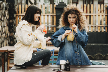 Portrait of two girlfriends wearing jeans blue and yellow jackets and drinking fresh juice.