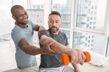 Favourite job. Delighted nice therapist smiling to his patient while helping his to lift a hand