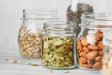 Various Nuts And Seeds In Glass Jars Over White Wooden Table Against White Background. The Concept Of Vegetarian And Organic Food. Set Of Photos.