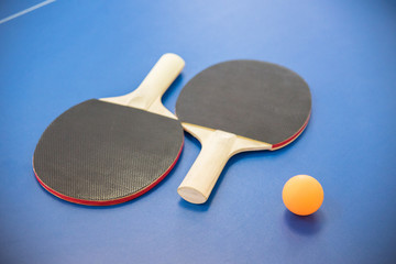 Orange ball for table tennis and two rackets of red and black color on a blue table with a grid