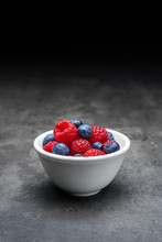 White Ceramic Bowl With Berry Fruit, Blueberries And Raspberries On Dark Concrete Background. Close Up With Copy Space At The Top.