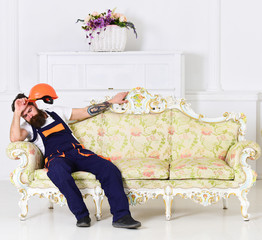 Wall Mural - Loader sit on sofa, having rest. Man with beard, worker in overalls and helmet sits on couch tired, white background. Exhausted loader concept. Courier relaxing while moving furniture, relocation.