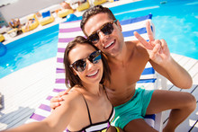 Self Portrait Of Attractive, Stylish, Cheerful Couple In Summer Glasses, Spectacles Showing Two Fingers Shooting Selfie On Front Camera Sitting On Lounger, Sunbed, Chaise-longue Near Swimming Pool