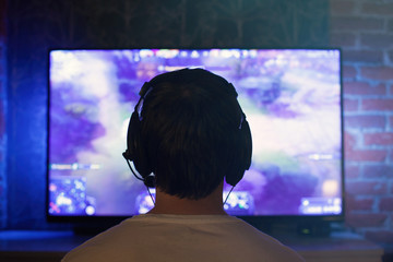 Gamer or streamer in earphones with microphone sits at home in dark room and plays with friends on networks in video games. A young man sits in front of a monitor or TV