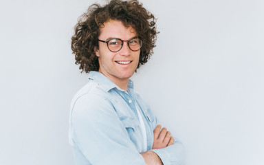 Wall Mural - Horizontal profile view portrait of handsome smiling young male with curly hair, wears denim shirt and round trendy spectacles, looking to the camera. Copy space for advertisement. People race concept