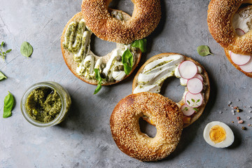 Variety of homemade bagels with sesame seeds, cream cheese, pesto sauce, eggs, radish, herbs served on crumpled paper with ingredients above over grey texture background. Top view, space.