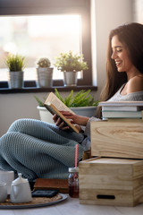 Wall Mural - Beautiful young woman reading a book, enjoying by the window in modern apartment. 