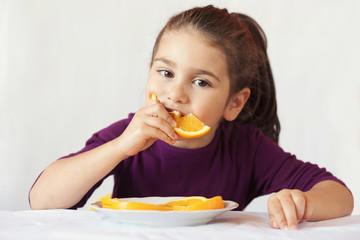 Wall Mural - little cute child girl dressed in a purple blouse holding an orange and eating off a piece of orange