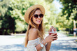Smiling attractive girl in sunglasses holds a fresh cocktail, looking at camera, wearing trendy clothes and hat. Summertime.
