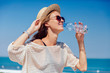 Summer photo of a happy pretty girl in sunglasses drinking clear water, during walking on quay, near the ocean. Dressed in stylish clothes and hat.