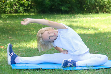 School-age happy kid girl in light clothes take sport exercise on a mat in the park. Outdoors workout. Healthy lifestyle. 