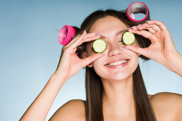 Poster - happy young girl smiling, holding slices of cucumbers near the face, on head of curlers