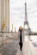 Young woman in grey coat standing on Trocadero square near gilded statues and Eiffel Tower in Paris. Concept of trip to France and European landmarks.