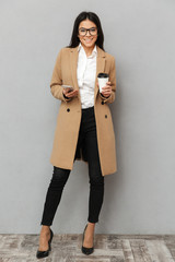 full length image of beautiful woman wearing outerwear standing holding with mobile phone and takeaw