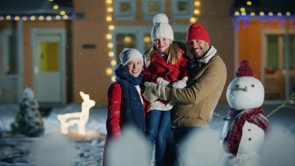 Wall Mural - Happy Young Family Portrait in the Falling Snow, Father Holds  Daughter and Embraces His Wife. Smiling Family Enjoys Winter Holiday in the Front Yard of their Idyllic House Decorated with Garlands.