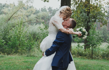 Wedding couple is hugging in the green forest. Bride in satin dress with train and tulle veil is holding a bouquet and smiling. High green trees and bright emerald green grass outdoors. Warm embrace.