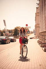 Wall Mural - Girl in a hat on a city bicycle with a basket, rides down the city street on a sunny day looking at the camera.