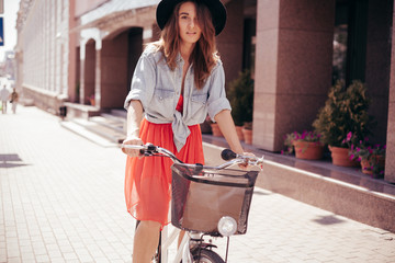 Wall Mural - Girl in a hat on a city bicycle with a basket, rides down the city street on a sunny day looking at the camera.