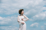 Fototapeta Konie - Girl on sunny blue sky background in summer. Beauty and fashion look in vintage style. Makeup on young face. Fashion woman model pose outdoor. Pretty girl in white wedding dress and retro hairstyle.