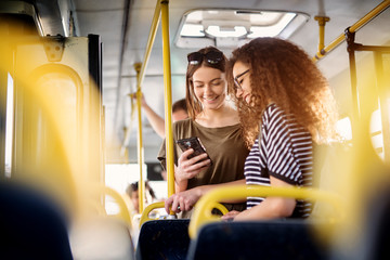 two cheerful pretty young women are standing in a bus and looking at the phone and smiling while wai