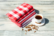White  Cup Of Hot Black Coffee With Roasted Coffee Beans Together With A Stack Of Red White Checkered And Striped Linen Tableclothes On Rustic Bright Wooden Table. Outdoor.Card Concept.