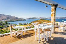Your Table Is Already Reserved At The Greek Mediterranean Sea Behind Crystal Clear Blue Water And A Beautiful Coastal Aegean Bay With Fishermen Boats Cruising, Patmos Island, Kos, Dodecanese, Greece