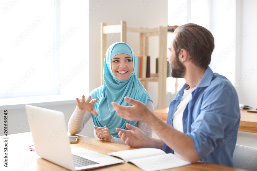 Male student and his Muslim classmate using laptop in library
