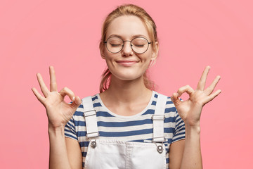 Wall Mural - Portrait of satisfied blonde attractive delighted female indicates ok sign, keeps eyes shut, being in good mood, happy to have weekends, isolated over pink background. Student girl gestures.