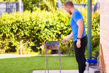 Handsome Athlete Male Prepares Barbecue Outdoors. Brutal Bearded Man Standing And Prepared Grill, Burn Charcoal Outdoor With Sunset Nature Summer Garden Background.