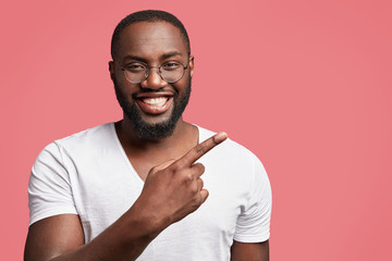 Wall Mural - Attractive positive dark skinned man with broad smile, indicates at blank copy space of pink background for your advertising content or promotional text. Happy unshaven male seller of goods.