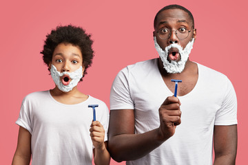 Surprised dark skinned father and son with applied shaving gel on faces, hold razors with stunned expressions, have fun together, learn how to shave, isolated over pink background. Hygiene concept