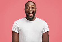 Waist Up Portrait Of Glad Dark Skinned Handsome Male Model With Happy Expression, Wears Round Spectacles, Being In Good Mood As Recieve Bonus For Diligent Work, Isolated Over Pink Background