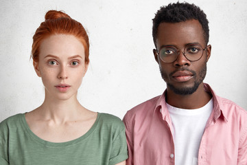 Wall Mural - Young freckled Caucasian girl with ginger hair and her best African American friend, look directly at camera with serious expression, listen important information. Dark skinned male student in eyewear